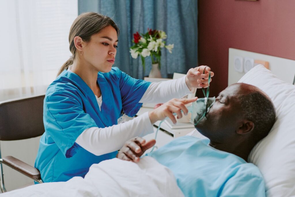 A nurse assists a bedridden patient with oxygen support in a hospital room.