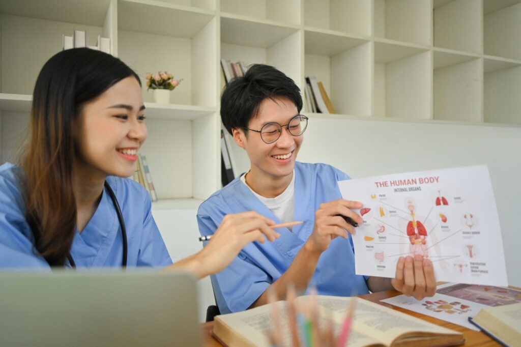 Two cheerful nurses review the human anatomy poster with open textbooks spread across a study table.