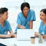 Three nurses in blue scrubs gathered around a laptop, discussing the study materials in a bright break room.