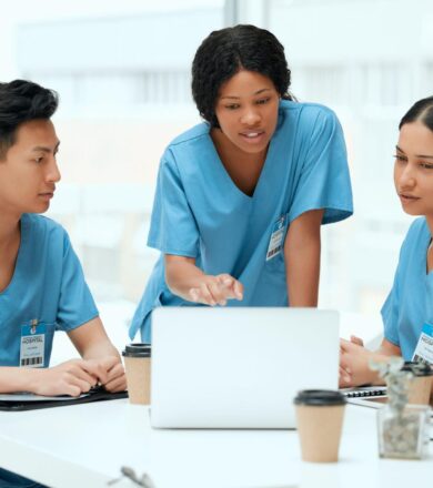 Three nurses in blue scrubs gathered around a laptop, discussing the study materials in a bright break room.