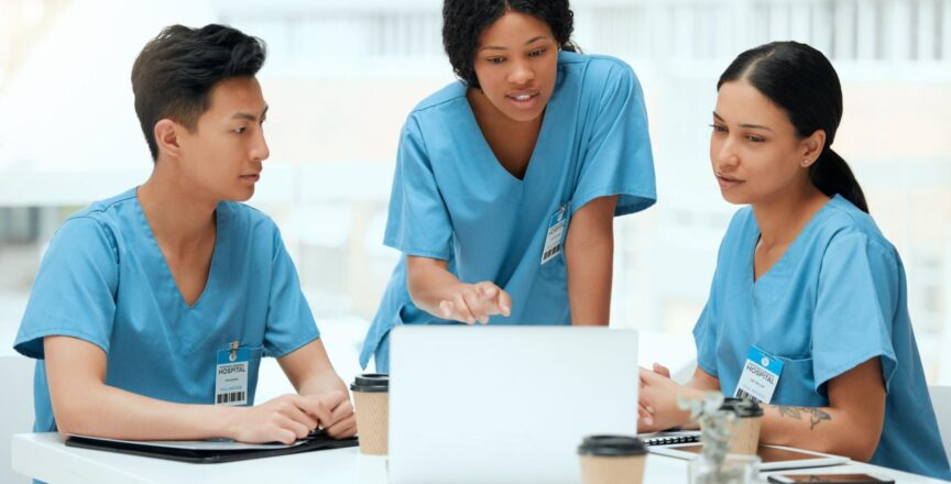 Three nurses in blue scrubs gathered around a laptop, discussing the study materials in a bright break room.
